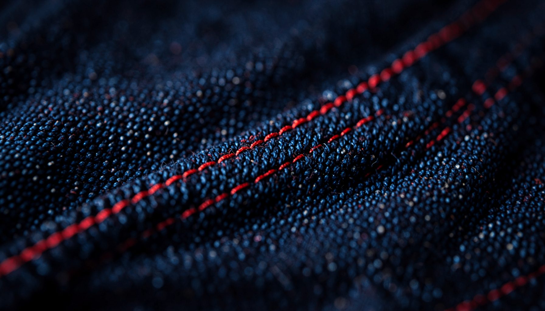 Ảnh cận cảnh chi tiết bề mặt vải denim xanh đậm với các đường chỉ đỏ nổi bật trên nền tối, tạo cảm giác ấm áp và công nghiệp.